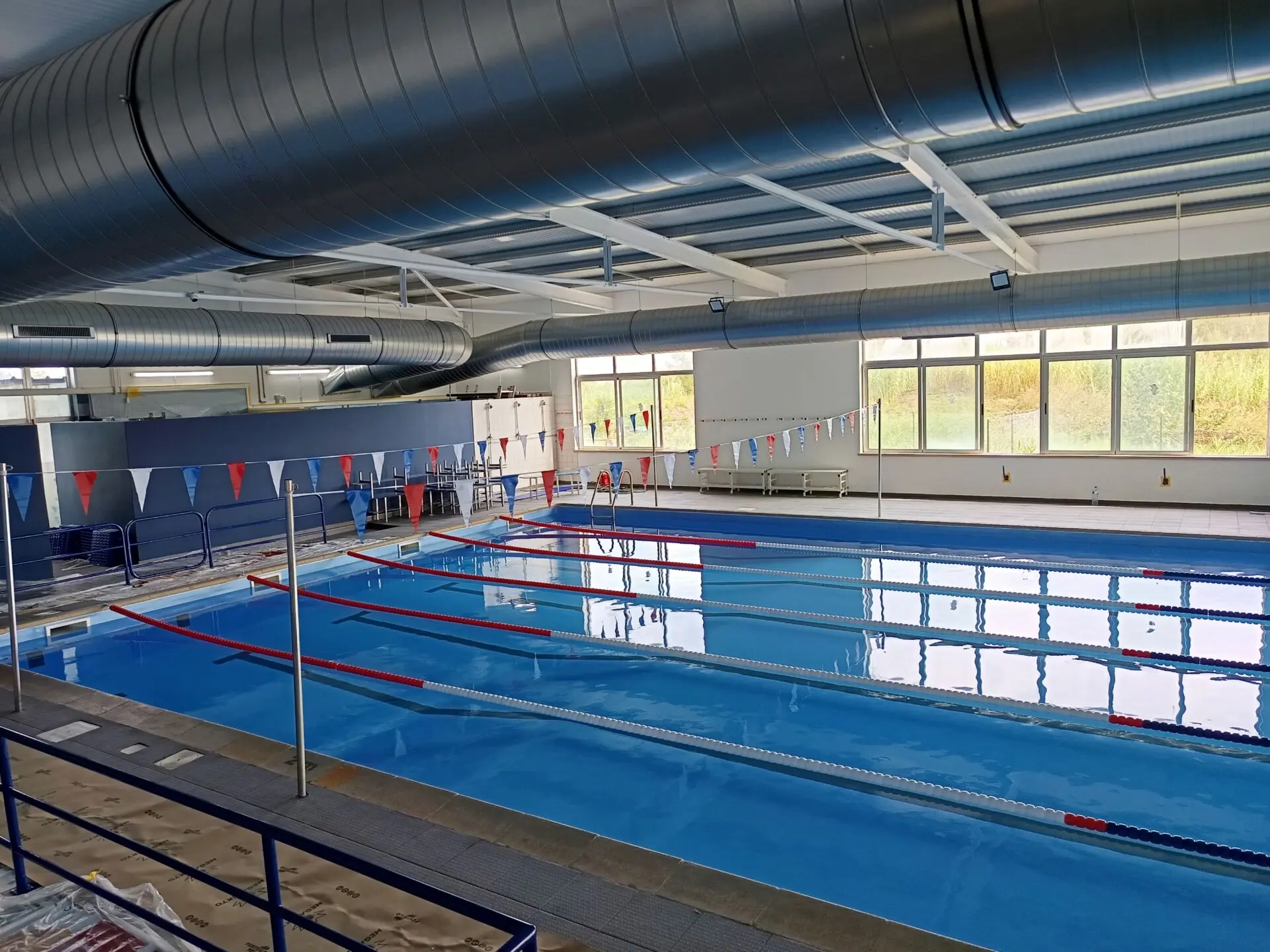 A imagem mostra uma piscina interior vazia, com pistas delimitadas por separadores e bandeirolas suspensas. São visíveis janelas amplas e condutas de ventilação no teto.