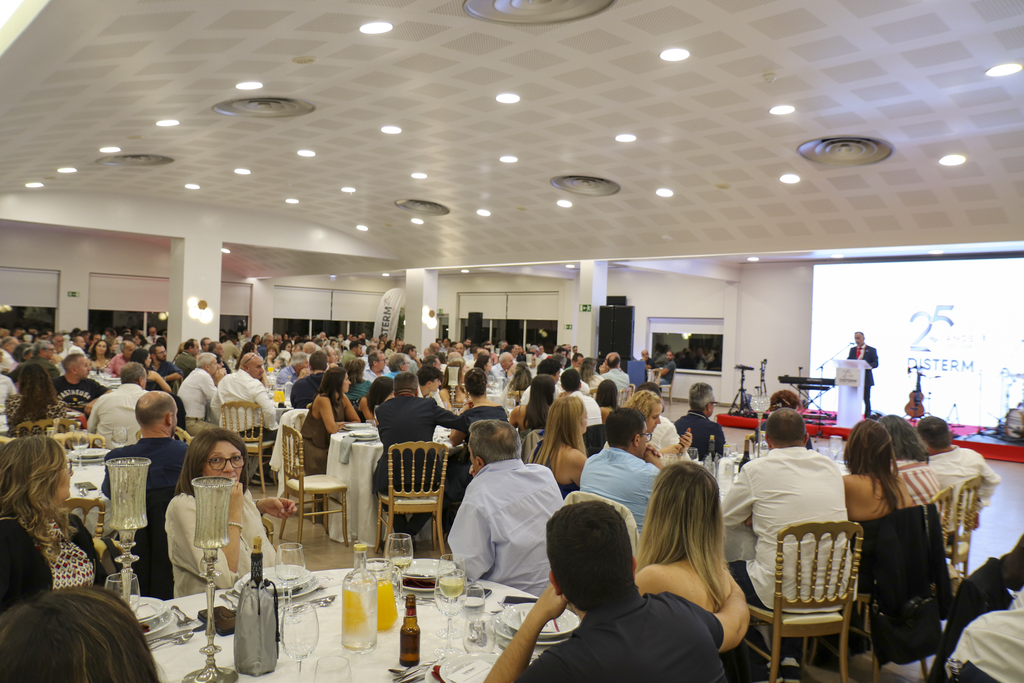 Um grande grupo de pessoas está reunido à mesa num salão durante um jantar. À direita, um orador discursa num palco junto a uma projeção onde se lê «25» e «PISTERM».