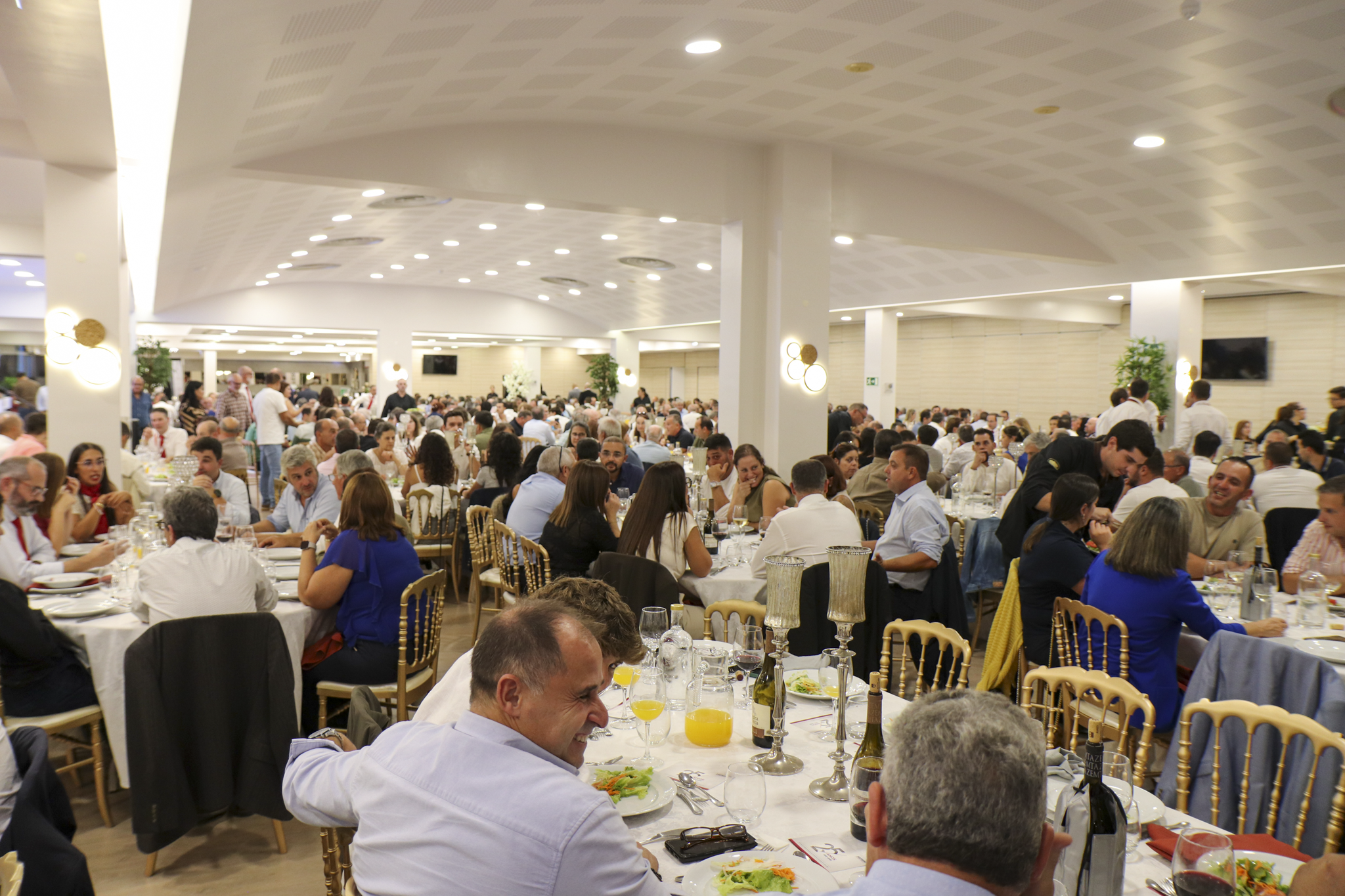 Um grande grupo de pessoas está reunido para uma refeição num salão de banquetes. As mesas estão postas com pratos, copos e bebidas.