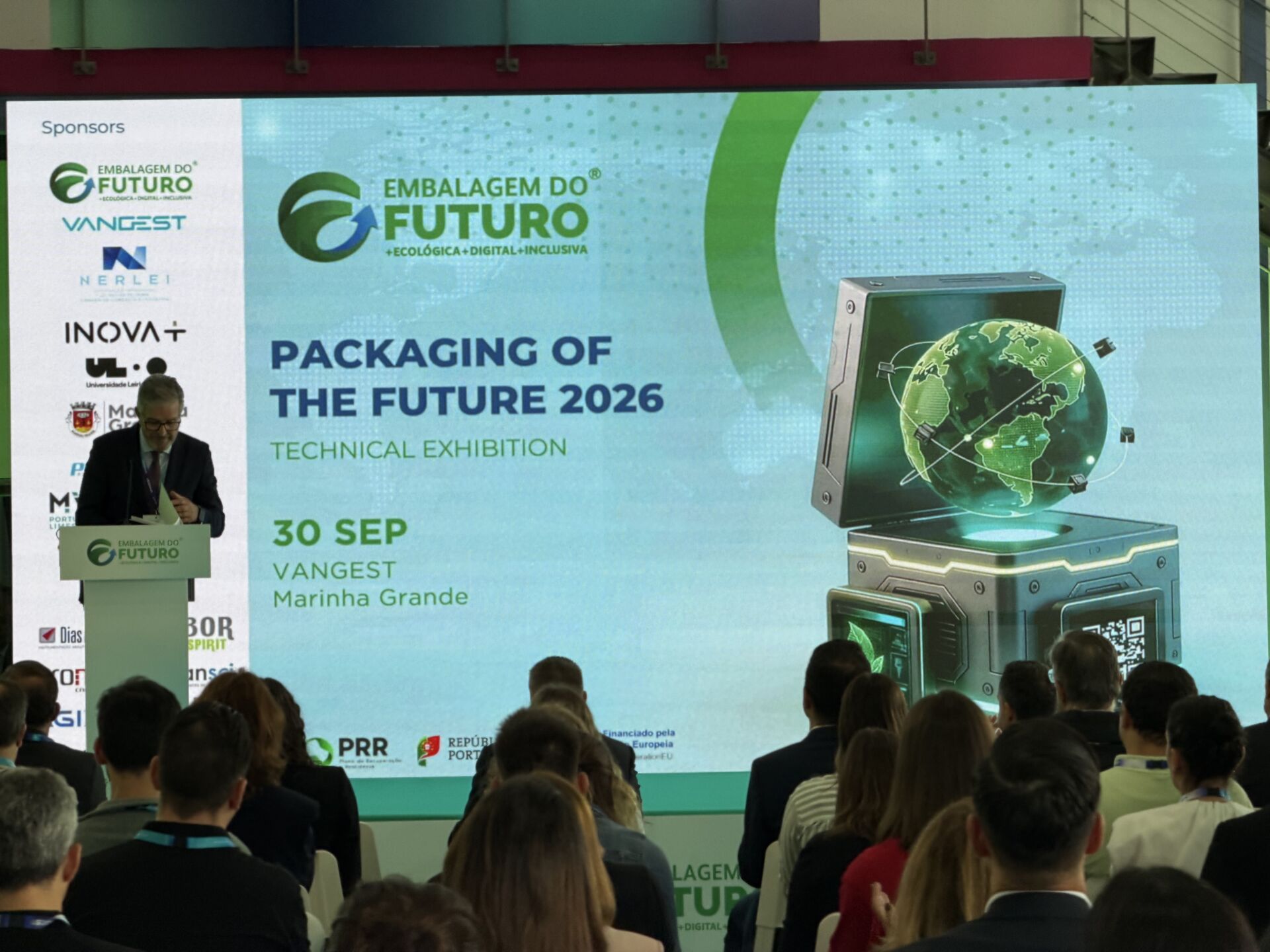 Um orador discursa perante uma plateia, junto a um ecrã que anuncia a exposição técnica «Packaging of the Future 2026», em 30 de setembro, na Vangest, Marinha Grande. São visíveis os logótipos de várias entidades patrocinadoras.