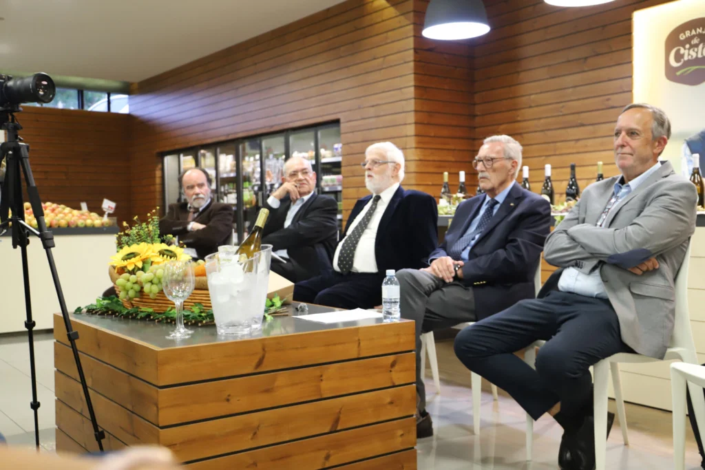 Cinco homens estão sentados lado a lado num espaço comercial, junto a uma mesa com fruta, flores e bebidas. À esquerda, vê-se uma câmara montada num tripé.