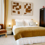 Quarto com uma cama de casal, uma colcha em tons de ocre e duas gravuras abstratas na parede. À esquerda, há um cadeirão junto a uma mesa de cabeceira com um candeeiro; à direita, um biombo escuro.