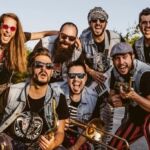 Sete músicos posam ao ar livre, alguns com instrumentos de sopro e uma guitarra. Vestem coletes de ganga e olham para a câmara.