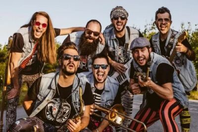 Sete músicos posam ao ar livre, alguns com instrumentos de sopro e uma guitarra. Vestem coletes de ganga e olham para a câmara.