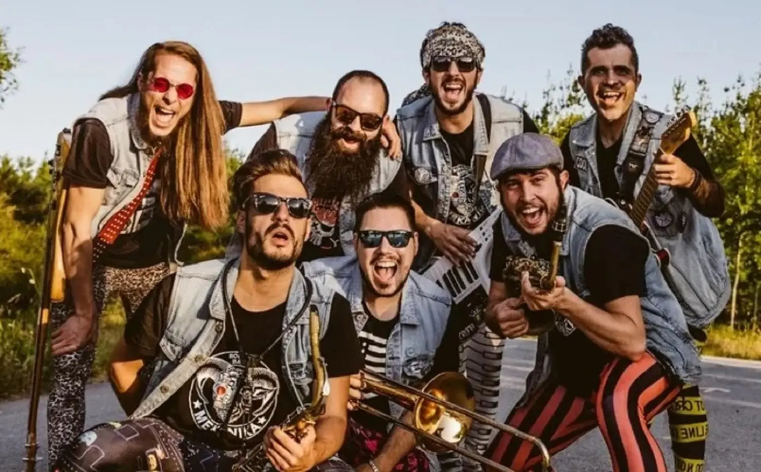 Sete músicos posam ao ar livre, alguns com instrumentos de sopro e uma guitarra. Vestem coletes de ganga e olham para a câmara.