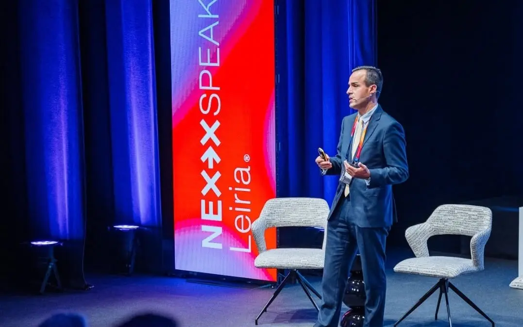 Um homem de fato apresenta-se em palco, com microfone e comando na mão. Ao fundo, vê-se um painel com a inscrição «NEXX SPEAKER» e «Leiria».