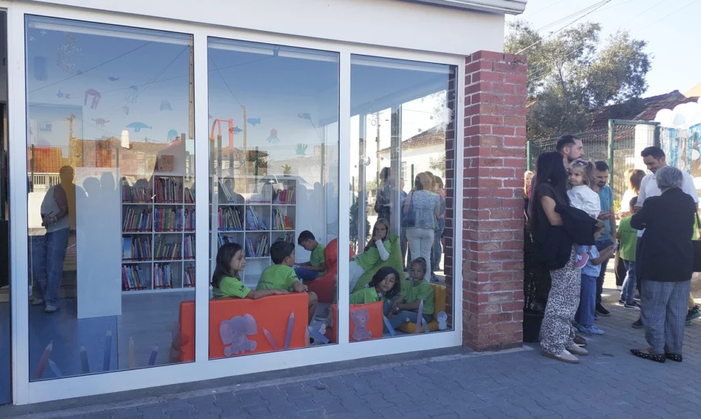 Crianças estão sentadas numa sala envidraçada com estantes de livros, enquanto um grupo de adultos conversa no exterior. A imagem mostra um espaço de leitura infantil.