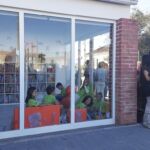 Crianças estão sentadas numa sala envidraçada com estantes de livros, enquanto um grupo de adultos conversa no exterior. A imagem mostra um espaço de leitura infantil.