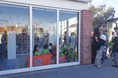 Crianças estão sentadas numa sala envidraçada com estantes de livros, enquanto um grupo de adultos conversa no exterior. A imagem mostra um espaço de leitura infantil.