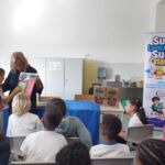 Uma mulher apresenta um folheto a um grupo de crianças, com outro adulto presente. Um cartaz identifica a atividade como parte do projeto de educação alimentar «Super Lancheiras, Super Crianças», em escolas do 1.º ciclo do concelho de Leiria.