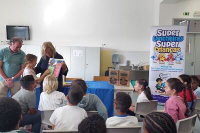 Uma mulher apresenta um folheto a um grupo de crianças, com outro adulto presente. Um cartaz identifica a atividade como parte do projeto de educação alimentar «Super Lancheiras, Super Crianças», em escolas do 1.º ciclo do concelho de Leiria.