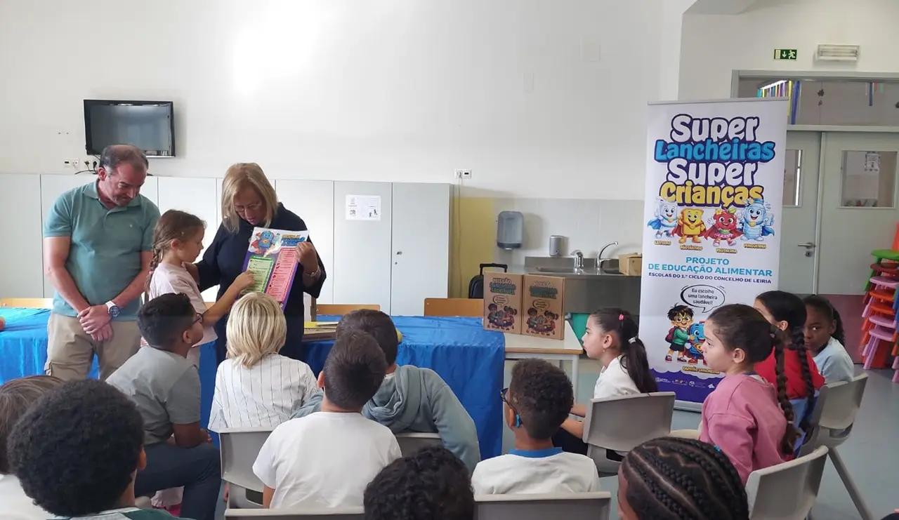 Uma mulher apresenta um folheto a um grupo de crianças, com outro adulto presente. Um cartaz identifica a atividade como parte do projeto de educação alimentar «Super Lancheiras, Super Crianças», em escolas do 1.º ciclo do concelho de Leiria.