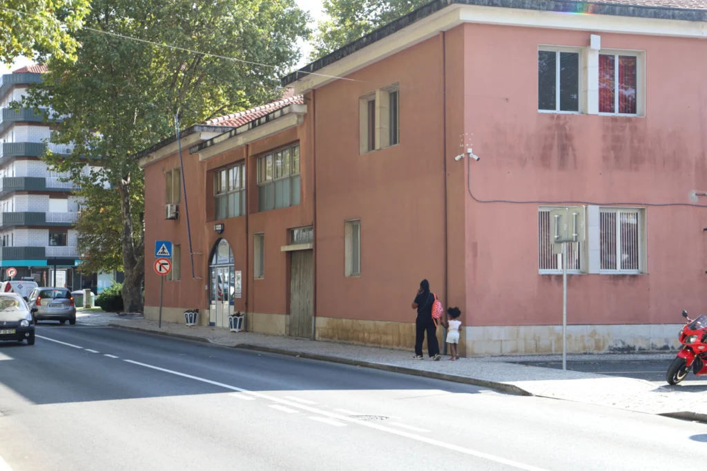 Uma rua com um edifício cor-de-rosa junto ao passeio. Uma pessoa adulta e uma criança caminham ao lado do edifício; veem-se carros e uma mota.