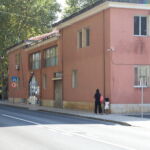 Uma rua com um edifício cor-de-rosa junto ao passeio. Uma pessoa adulta e uma criança caminham ao lado do edifício; veem-se carros e uma mota.