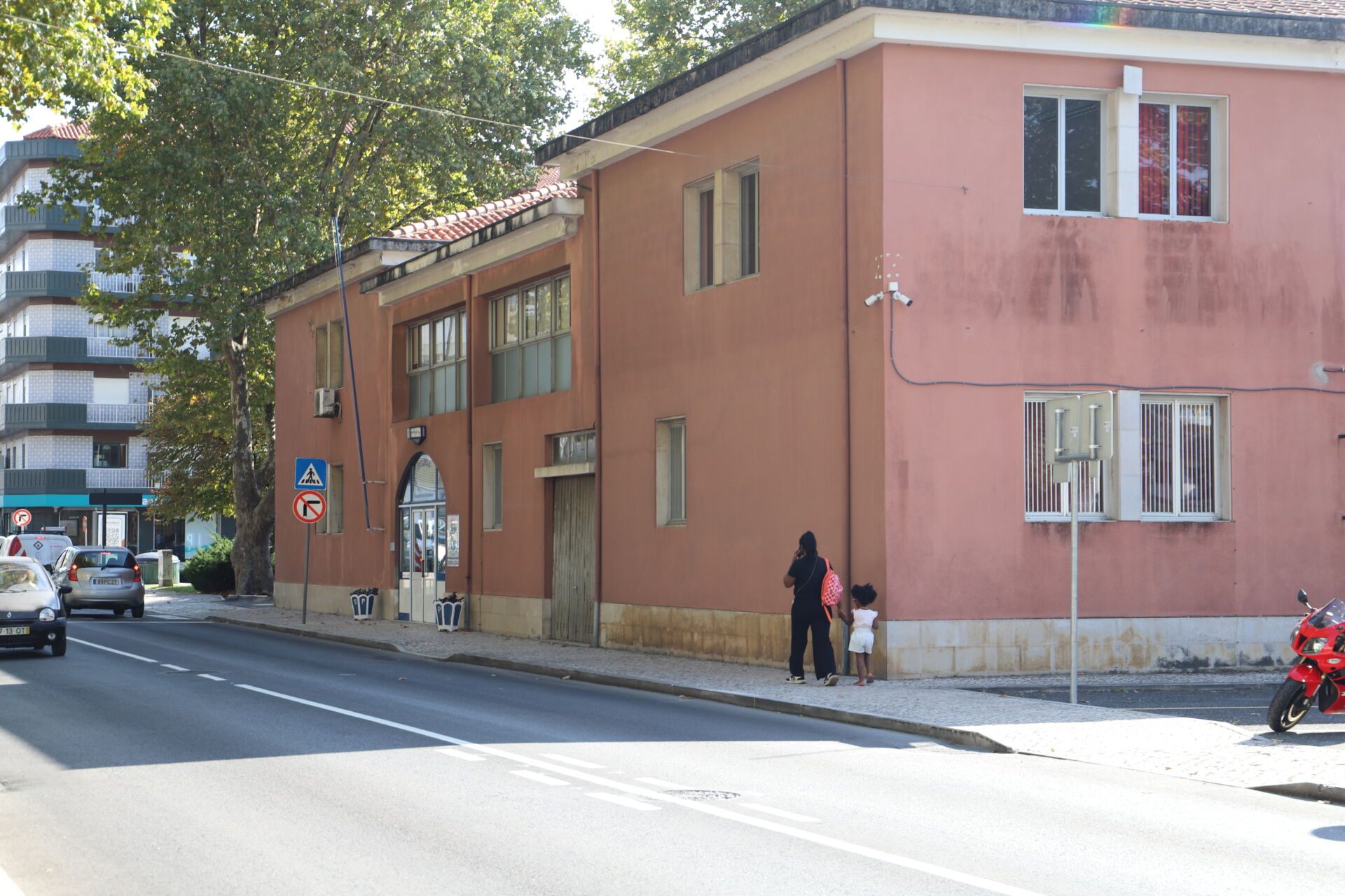 Uma rua com um edifício cor-de-rosa junto ao passeio. Uma pessoa adulta e uma criança caminham ao lado do edifício; veem-se carros e uma mota.