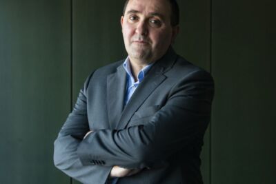 Retrato de um homem de fato escuro e camisola azul, de braços cruzados, diante de uma parede verde-escura. Não há texto visível que permita identificá-lo.