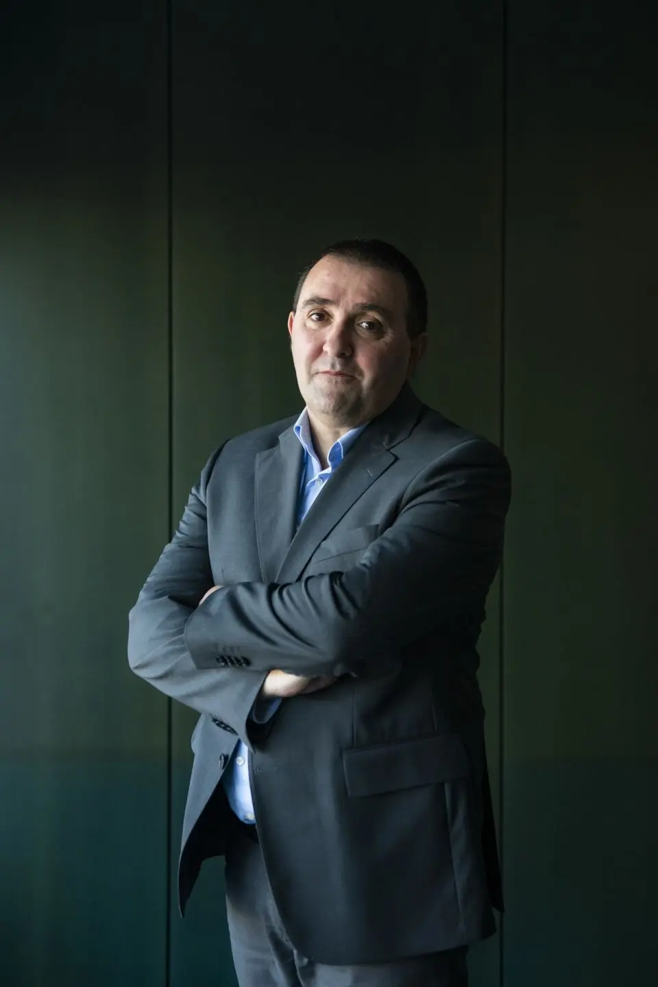 Retrato de um homem de fato escuro e camisola azul, de braços cruzados, diante de uma parede verde-escura. Não há texto visível que permita identificá-lo.