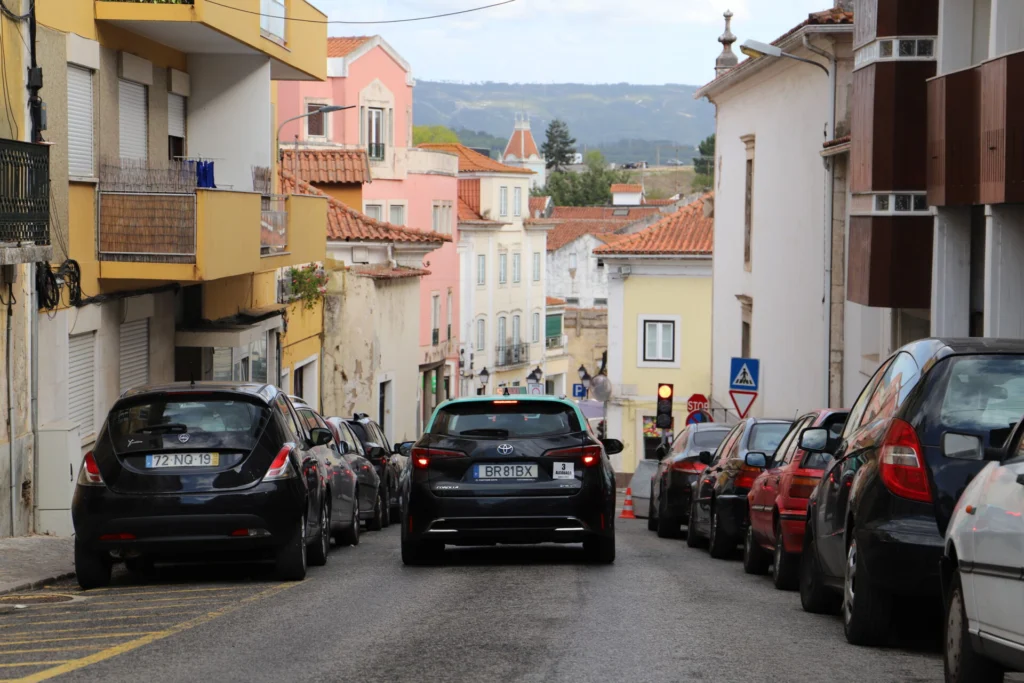 Uma rua urbana em declive, com carros estacionados de ambos os lados e um táxi a circular ao centro. Veem-se edifícios com telhados de telha e sinais de trânsito.