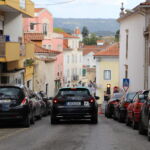 Uma rua urbana em declive, com carros estacionados de ambos os lados e um táxi a circular ao centro. Veem-se edifícios com telhados de telha e sinais de trânsito.