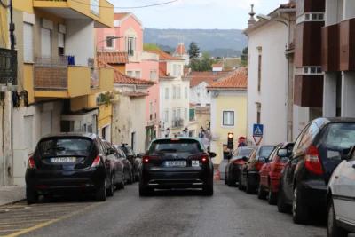 Uma rua urbana em declive, com carros estacionados de ambos os lados e um táxi a circular ao centro. Veem-se edifícios com telhados de telha e sinais de trânsito.