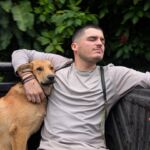 Um homem está sentado ao ar livre com o braço à volta de um cão castanho. Atrás deles vê-se vegetação densa.