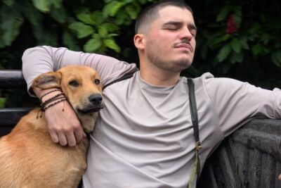 Um homem está sentado ao ar livre com o braço à volta de um cão castanho. Atrás deles vê-se vegetação densa.