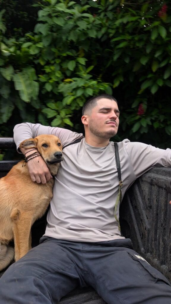 Um homem está sentado ao ar livre com o braço à volta de um cão castanho. Atrás deles vê-se vegetação densa.