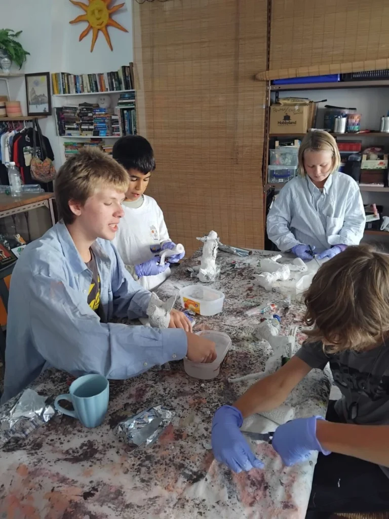 Quatro jovens trabalham em peças de modelagem à volta de uma mesa, usando luvas de proteção. O espaço tem livros, materiais e utensílios de trabalho manual.