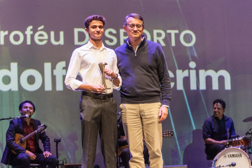 Dois homens posam em palco; um deles segura um troféu. Ao fundo, vêem-se músicos e texto projetado parcialmente visível.