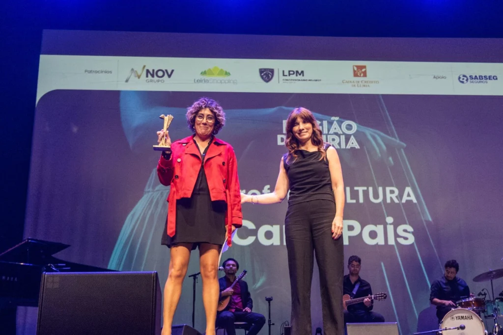 Duas mulheres posam em palco durante uma cerimónia ligada à cultura; uma delas segura um troféu. Ao fundo, vêem-se músicos e os logótipos dos patrocinadores.