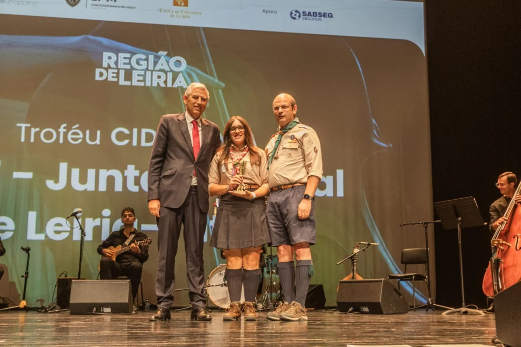 Três pessoas posam no palco, uma delas a segurar um troféu, perante uma projeção com as inscrições «Região de Leiria» e «Troféu CID». Dois dos participantes vestem uniformes de escuteiros; há músicos ao fundo.