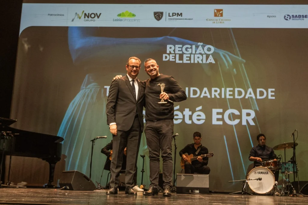 Dois homens posam num palco, um deles com um troféu. Ao fundo, uma projeção da Região de Leiria destaca a palavra «Solidariedade», com músicos sentados atrás.