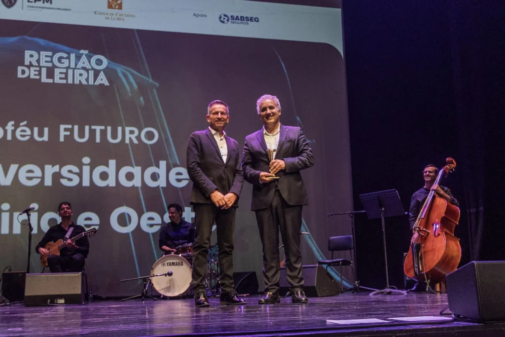 Dois homens posam em palco, um deles com um troféu, durante um evento associado à Região de Leiria. Ao fundo, vêem-se músicos e uma projeção com texto parcialmente cortado sobre o futuro e a diversidade.