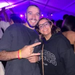 Duas pessoas sorriem e posam para a fotografia num espaço coberto, com outras pessoas ao fundo. A iluminação é predominantemente roxa.