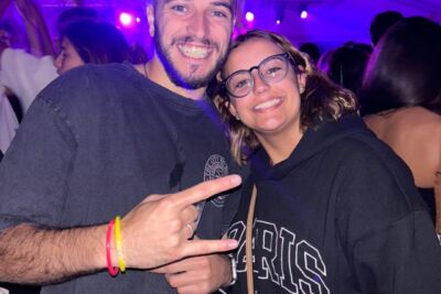 Duas pessoas sorriem e posam para a fotografia num espaço coberto, com outras pessoas ao fundo. A iluminação é predominantemente roxa.