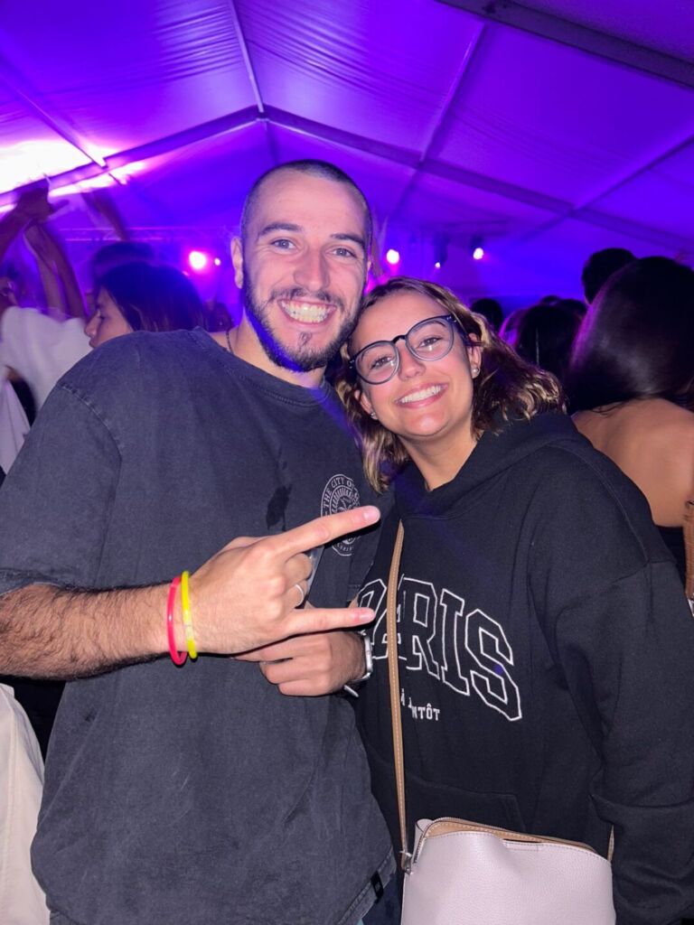 Duas pessoas sorriem e posam para a fotografia num espaço coberto, com outras pessoas ao fundo. A iluminação é predominantemente roxa.