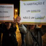 Duas pessoas seguram cartazes numa manifestação noturna pelo direito à habitação. Os cartazes afirmam que as casas são para viver, não para enriquecer, e que a habitação não é um luxo.