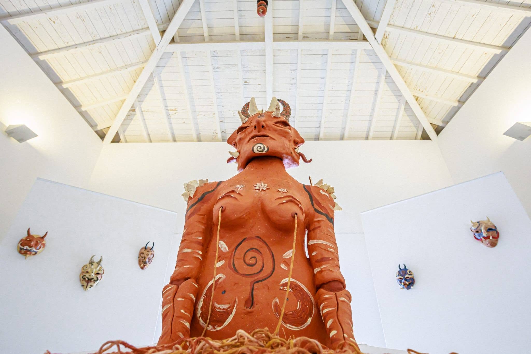Uma grande figura escultórica avermelhada, com cornos e motivos em espiral, ocupa o centro de uma sala de paredes brancas. Nas paredes estão expostas várias máscaras coloridas.