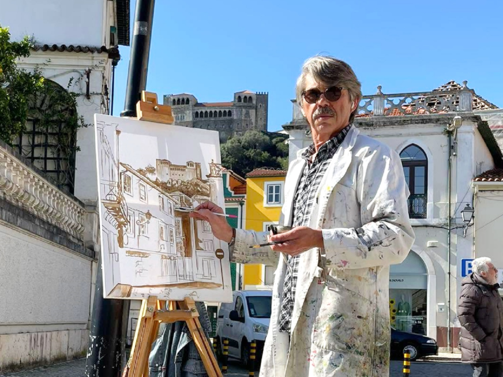 Um homem pinta uma paisagem urbana num cavalete, ao ar livre. Ao fundo veem-se edifícios e um castelo.