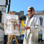 Um homem pinta uma paisagem urbana num cavalete, ao ar livre. Ao fundo veem-se edifícios e um castelo.