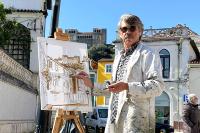 Um homem pinta uma paisagem urbana num cavalete, ao ar livre. Ao fundo veem-se edifícios e um castelo.