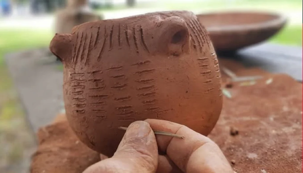Uma mão segura uma pequena peça de barro com marcas incisas, enquanto segura também um instrumento fino. Ao fundo, veem-se outras peças de barro sobre uma mesa.