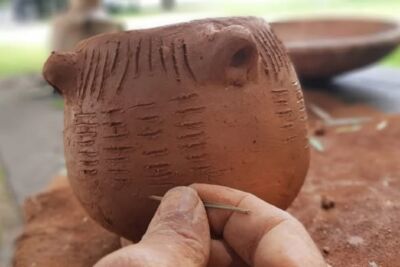 Uma mão segura uma pequena peça de barro com marcas incisas, enquanto segura também um instrumento fino. Ao fundo, veem-se outras peças de barro sobre uma mesa.