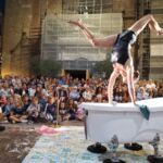 Uma artista faz uma acrobacia em equilíbrio sobre uma banheira, perante uma multidão, num espetáculo de rua noturno. O edifício ao fundo está coberto por andaimes.