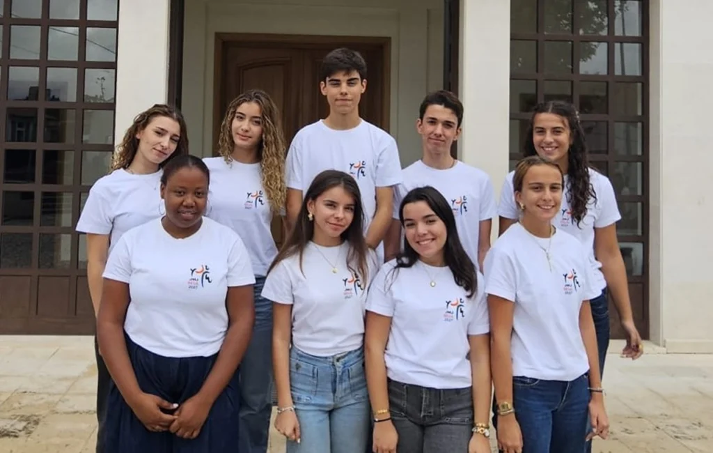 Nove jovens posam em grupo à entrada de um edifício. Vestem t-shirts brancas com o mesmo logótipo.