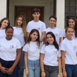Nove jovens posam em grupo à entrada de um edifício. Vestem t-shirts brancas com o mesmo logótipo.