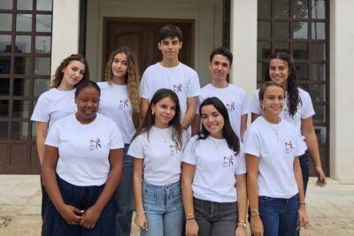 Nove jovens posam em grupo à entrada de um edifício. Vestem t-shirts brancas com o mesmo logótipo.