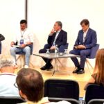Cinco intervenientes participam num painel de debate perante uma audiência. Um deles segura um microfone; há mesas com água à frente do painel.
