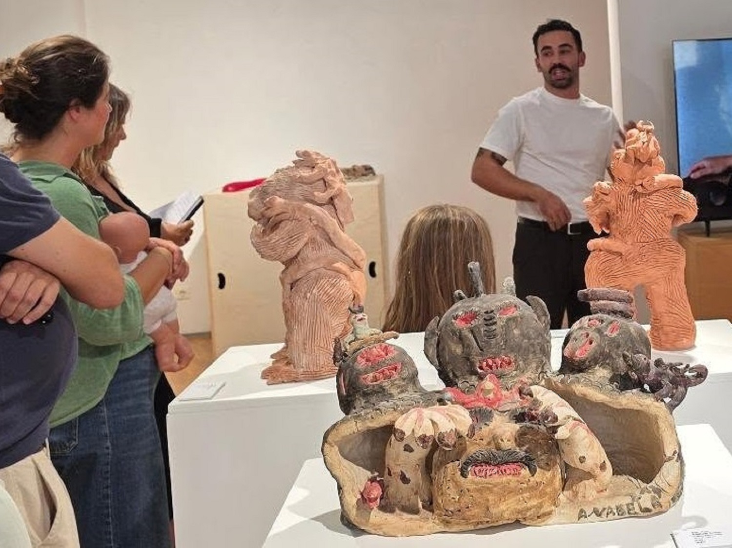 Um homem fala a um pequeno grupo junto de várias esculturas expostas numa sala. Em primeiro plano, vê-se uma peça com figuras e rostos; atrás, há duas esculturas de tom rosado.