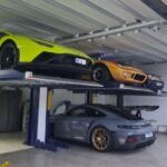 Numa garagem, vários automóveis desportivos estão estacionados em plataformas elevatórias. Um automóvel cinzento encontra-se por baixo das plataformas.